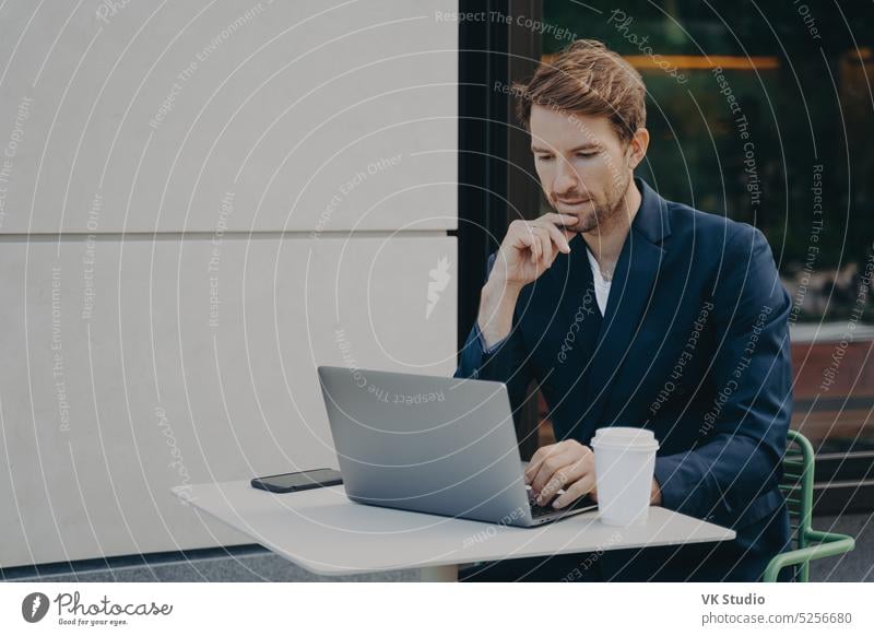 Gut aussehender, nachdenklicher junger Mann, der im Internet auf einem Laptop surft, während er in einem Straßencafé arbeitet Geschäftsmann Café Kaffee Tisch