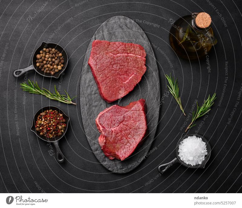 Rohes Stück Rindfleisch mit Gewürzen Pfeffer, Rosmarinzweig, Salz und Olivenöl auf einem Holzbrett, schwarzer Hintergrund. Fleisch niemand Paprika Vorbereitung