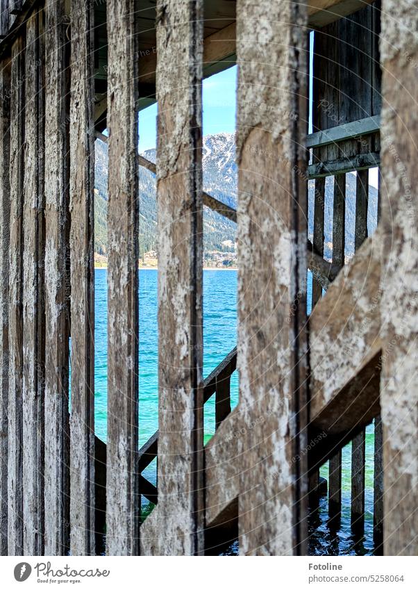 Durch die maroden Balken eines alten Bootsschuppens ist das türkisblaue Wasser des See's zu sehen und im Hintergrund türmen sich die Berge auf. Urlaub Gebirge