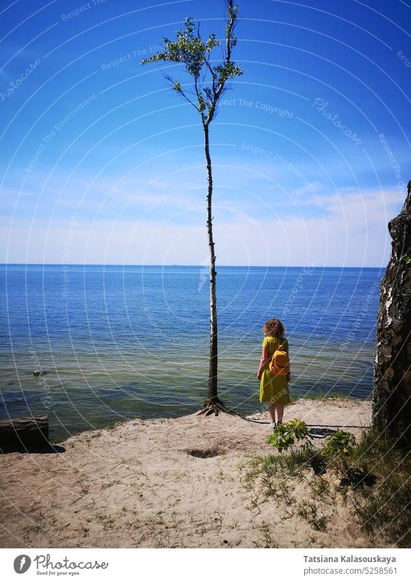 Eine lockige Frau in einem grünen Kleid mit einem gelben Rucksack steht am Ufer der Ostsee in Palanga und blickt von einem Steilufer aus in die Ferne zum Horizont. In der Nähe wächst ein einsamer Baum auf dem Sand.