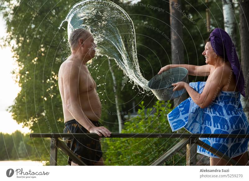Älteres Paar vergnügt sich nach finnischer Sauna auf einem Holzsteg in einem See. Ältere Frau gießt kaltes Wasser aus einem Becken über ihren Partner. Typischer finnischer Sommer.