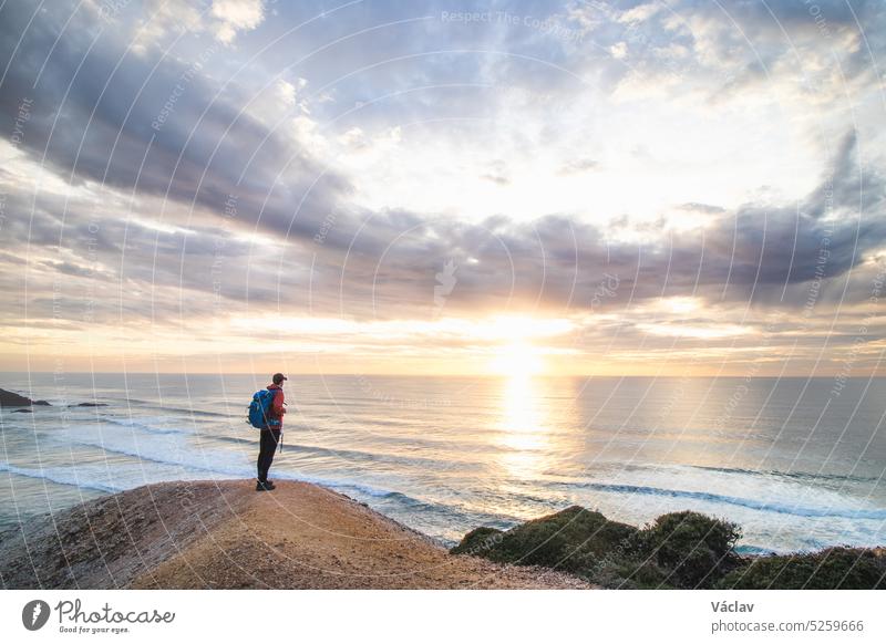 Ein Reisender steht am Rande einer Klippe in der Nähe der Stadt Odeceixe in der Region Algarve, Portugal, und beobachtet den Sonnenuntergang über dem klaren blauen Meer. Das Spiel der Farben am Himmel