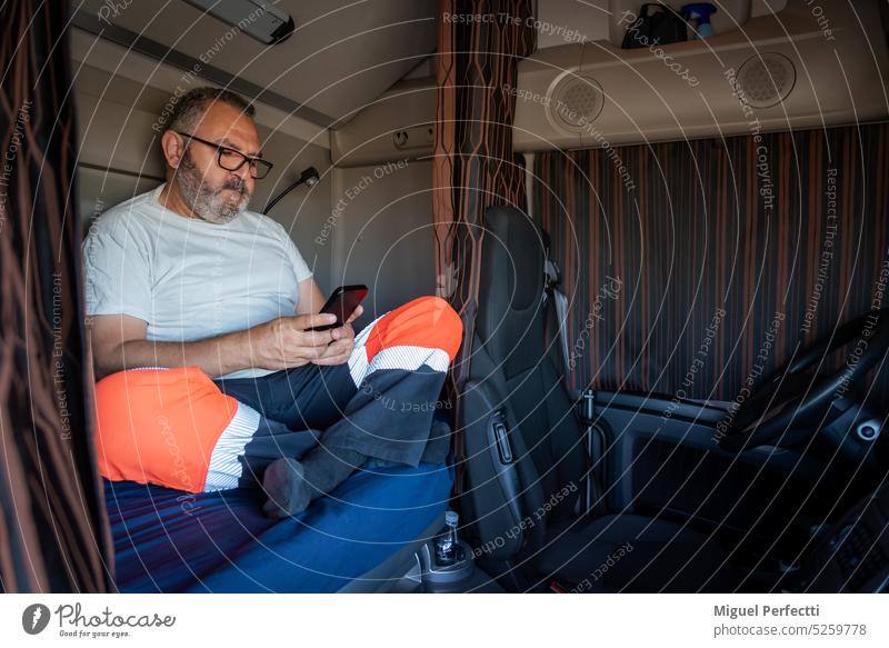 Ein älterer Lkw-Fahrer, der in seiner Freizeit bei zugezogenen Vorhängen auf der Ladefläche des Lkw sitzt und das Handy überprüft. Lastwagen Kabine Beruf Mann