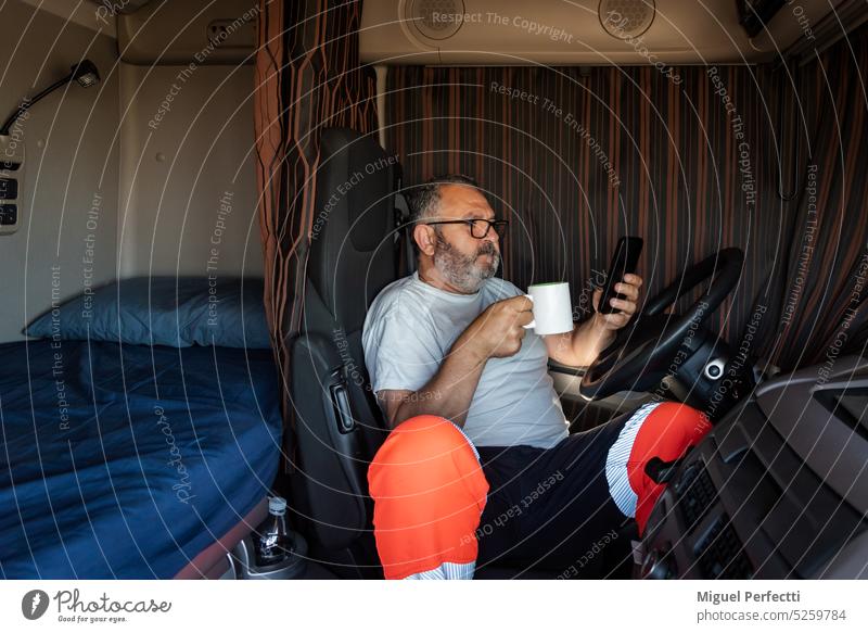 Ein älterer Trucker sitzt im Führerhaus seines Lastwagens, hat die Vorhänge zugezogen, trinkt seinen Morgenkaffee und schaut auf sein Telefon. Lkw-Fahrer Kabine