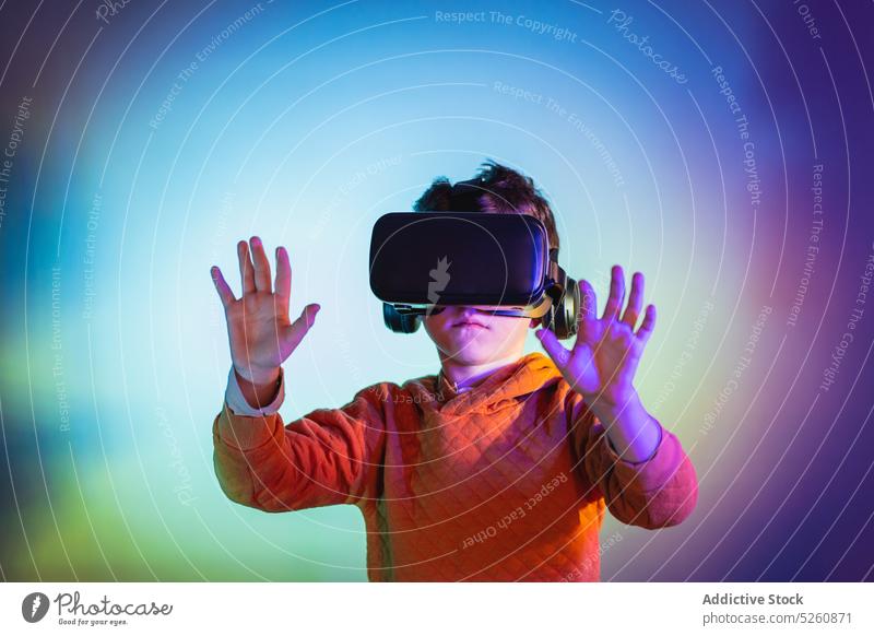 Junge erlebt VR-Headset-Spiel auf buntem Hintergrund Kind berühren unsichtbar Objekt Schutzbrille futuristisch Cyberspace Anschluss unterhalten