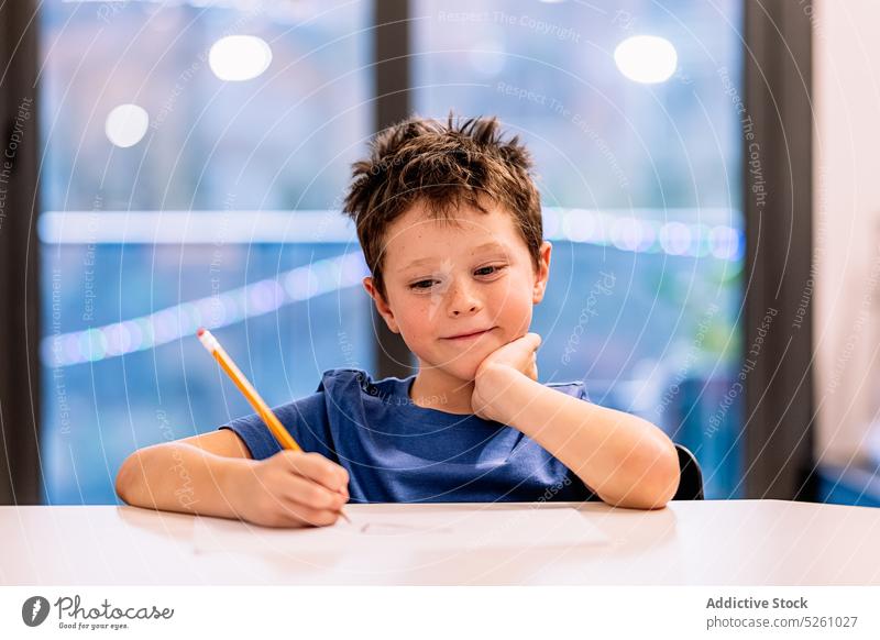 Fröhlicher Junge sitzt am Tisch und zeichnet zeichnen Lächeln heimwärts Fenster positiv kreativ Hobby Abend Kind sich auf die Hand lehnen Glück heiter froh