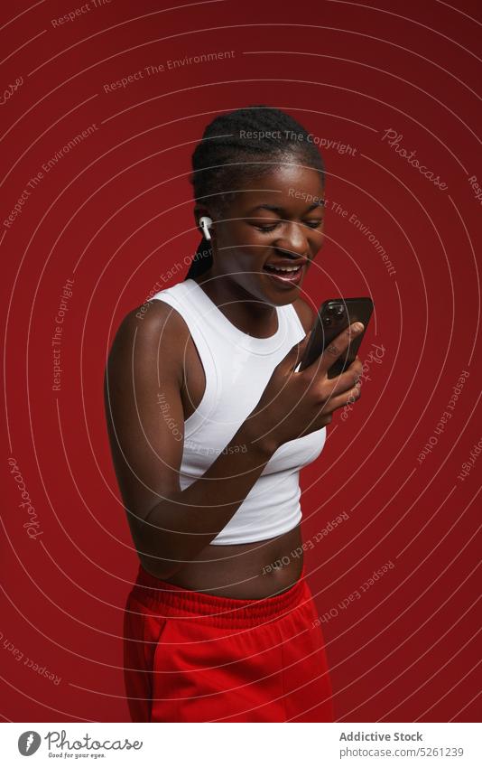 Fröhliche ethnische Frau in Sportkleidung mit Smartphone in der Nähe einer roten Wand heiter Glück zuhören Musik Kopfhörer benutzend Lächeln Model