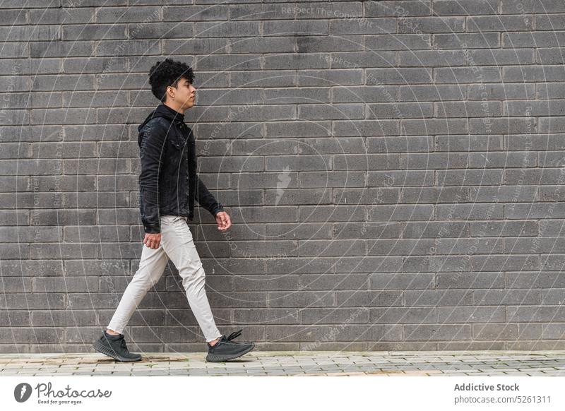 Junger männlicher Migrant geht auf der Straße in der Nähe einer Mauer Mann Schüler unabhängig Backsteinwand Gebäude einwandern Gelegenheit emotionslos Typ