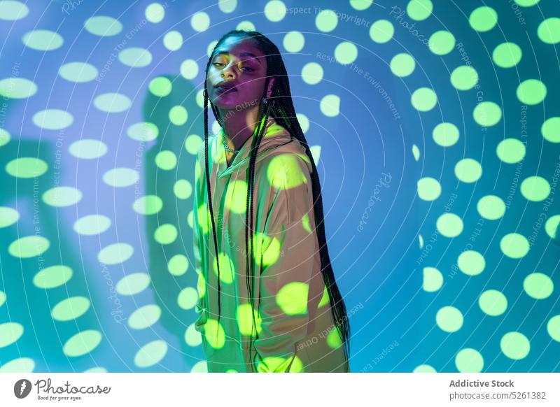 Schwarzes weibliches Modell, das vor einem leuchtenden Polka-Dot-Projektor steht Frau Punktmuster Muster neonfarbig Licht Einfluss emotionslos Afroamerikaner