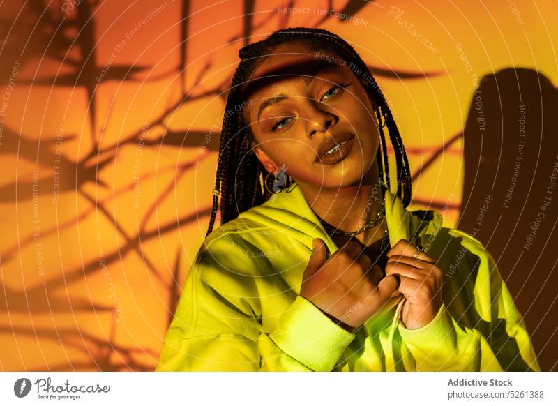 Schwarze Frau, die eine Kette berührt, steht in einem Studio gegen orangefarbenes Licht Model Porträt Geometrie Persönlichkeit neonfarbig selbstbewusst berühren
