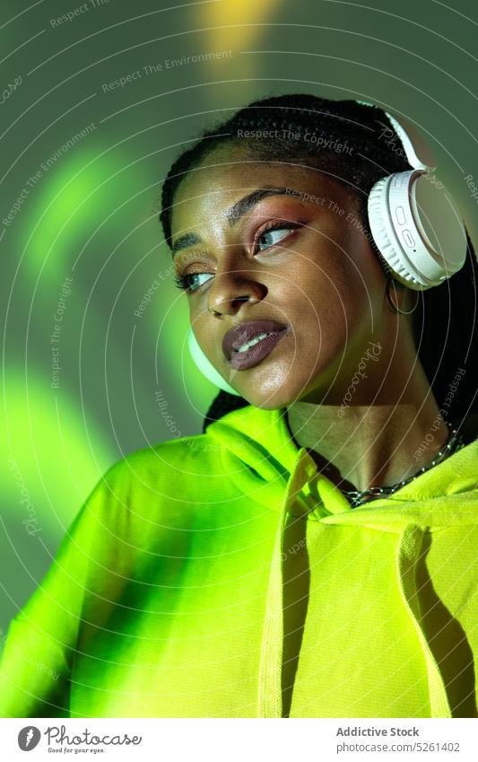 Schwarze Frau, die Kopfhörer anfasst, während sie in einem Studio mit Neonröhren steht Model berühren Klang zuhören Musik neonfarbig Licht Projektor