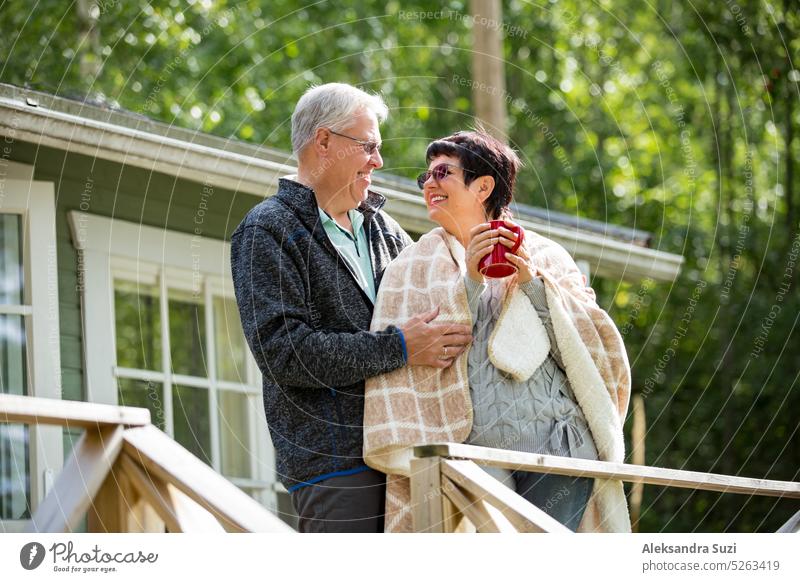 Ein älteres Paar steht auf einer Veranda und trinkt heißen Tee aus einer Tasse. Sie sind lächelnd und lachend, in eine Decke eingewickelt. Glückliches älteres Paar, das sich auf der Holzterrasse des Hauses umarmt.
