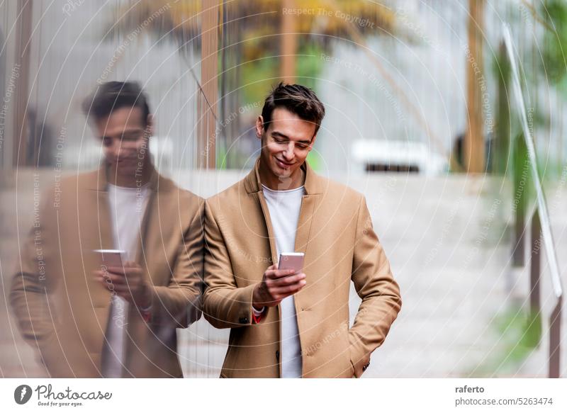 Porträt eines stilvollen, gut aussehenden jungen Mannes mit Mantel, der im Freien steht und sich an die Wand lehnt und ein Smartphone benutzt Erwachsener