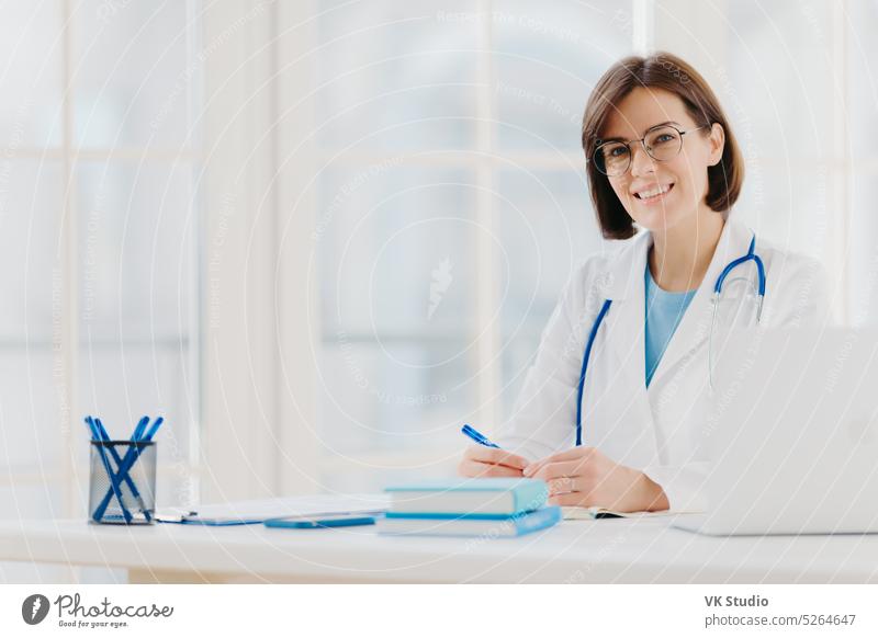 Medizinischer Spezialist trägt weißen Kittel mit Stethoskop, arbeitet im Krankenhaus, macht Notizen, posiert am eigenen Schrank, sitzt am Tisch mit Laptop im Büro und lächelt glücklich. Gesundheitspflege und medizinisches Konzept