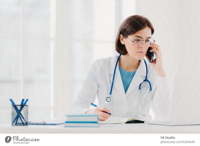 Gesundheitspersonal, Medizin-Konzept. Serious brunette weiblichen Arzt an modernen Laptop-Computer konzentriert, schreibt notwendige Informationen, spricht auf Handy, ruft jemand, hat ernsten Blick