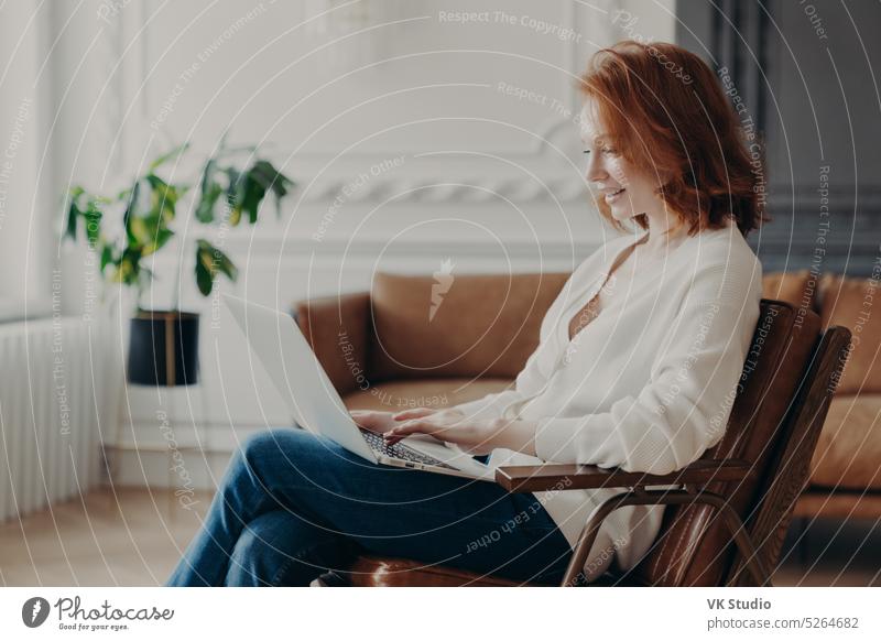 Lächelnde rothaarige Frau Freelancerin sitzt im Sessel, Tastaturen am Laptop-Computer, lädt notwendiges Material für die Entwicklung der Website hoch, posiert in moderner Wohnung, angeschlossen an drahtloses Internet.