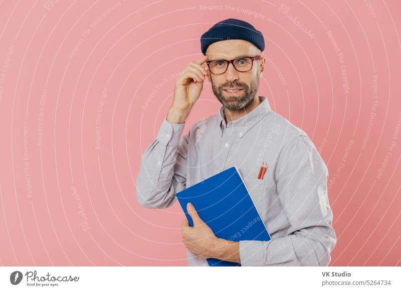 Stylish entfernten Arbeiter trägt transparente Brille, stilvolle Hut, Hemd, hält blauen Lehrbuch, verwendet Bleistifte für das Schreiben von Informationen, posiert über rosa Hintergrund. Zufriedene männliche Schriftsteller analysiert Informationen
