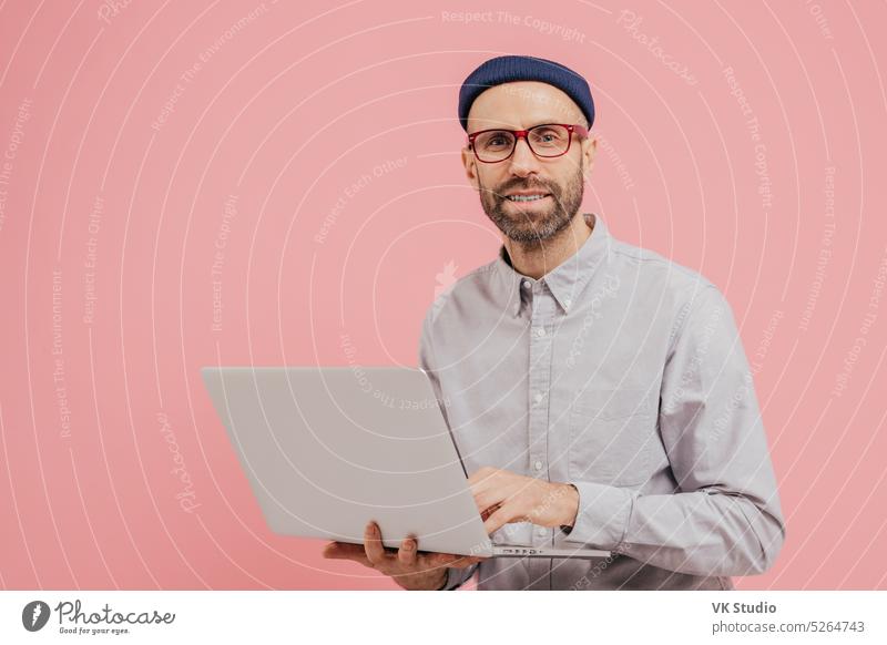 Foto von Kerl Internet-Nutzer surft Website auf Laptop-Computer, mit drahtlosem Internet verbunden, hält elektronische Gerät, trägt eine Brille, formale Hemd, isoliert über rosa Hintergrund mit freiem Raum