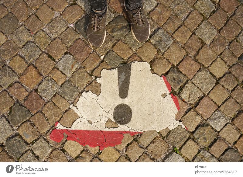 Blick von oben auf ein sich ablösendes Verkehrszeichen - Achtung Allgemeine Gefahrenstelle - , auf die Schuhe einer Person und auf altes Kopfsteinpflaster