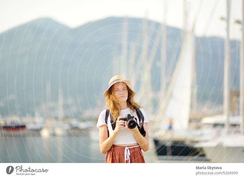 Charmante junge Touristin, die ein Foto an der Mittelmeerküste macht. Attraktive rothaarige Mädchen Fotograf mit einer Kamera auf dem Hintergrund von Meer und Yachten. Tourismus und Reisen