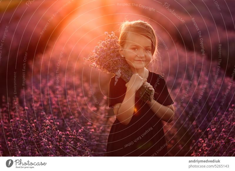 Porträt eines lächelnden niedlichen Mädchens mit einem Strauß Lavendelblüten in den Händen. Ein Kind geht in einem Feld von Lavendel auf Sonnenuntergang. Kind in schwarzem Kleid hat Spaß an der Natur im Sommerurlaub.