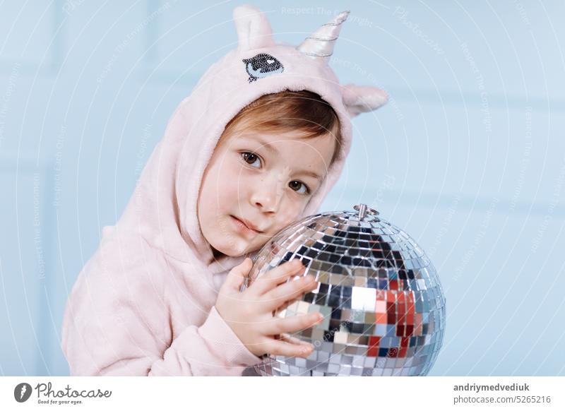 Lächelnde schöne kleine Mädchen in Einhorn rosa Kostüm lächelt und hat Spaß mit Discokugel auf einer blauen Wand Hintergrund. glückliche Kindheit. wenig Hut