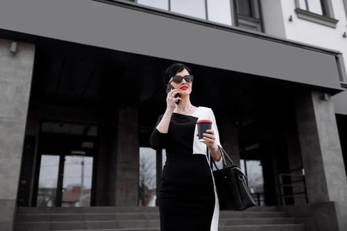 Erfolgreiche Geschäftsfrau Führer steht in großen Stadt spricht auf Handy. Lächelnde Frau, die geschäftliche Anrufe auf dem Handy zu Fuß mit einer Tasse Kaffee auf dem Hintergrund des Bürozentrums, Multitasking im Freien