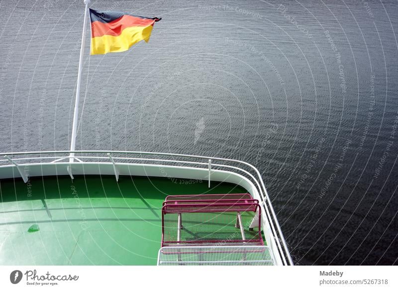Im Wind wehende deutsche Fahne in Schwarz, Rot und Gold bei Sonnenschein am Heck eines Ausflugsschiff auf dem Edersee bei Waldeck im Landkreis Waldeck-Frankenberg in Hessen