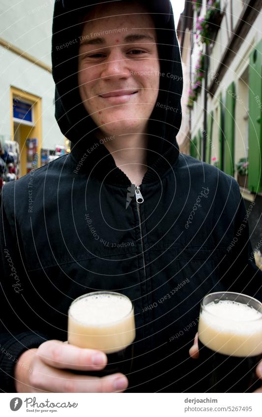 Ein junger Mann steht in Bamberg auf der Straße und hat zwei Gläser dunkle Biere in der Hand. Alkohol trinken Glas Getränk Schaum Außenaufnahme Durst Tag