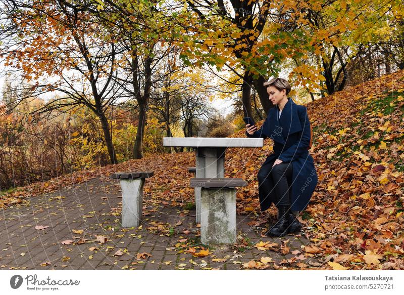 Eine Frau benutzt ein Smartphone, während sie an einem Tisch in einem herbstlichen Park sitzt fallen Herbst Telefon Erwachsener Kurze Haare Mobile blond