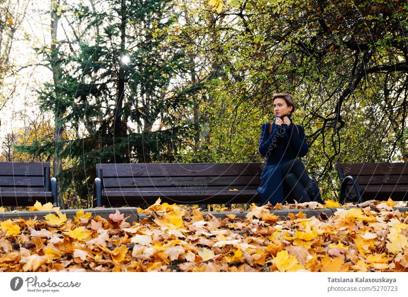 Eine Frau wickelt sich in einen Mantel ein und sitzt auf einer Bank in einem herbstlichen Park fallen Herbst Erwachsener Kurze Haare blond Kontemplation