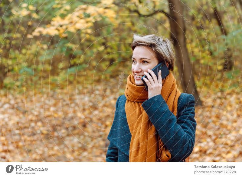 Kurzhaarige Blondine lächelt, als sie im Herbst im Freien telefoniert Frau fallen Anruf Telefon Mobile Handy Mitteilung am Telefon Blätter umgefallene Blätter