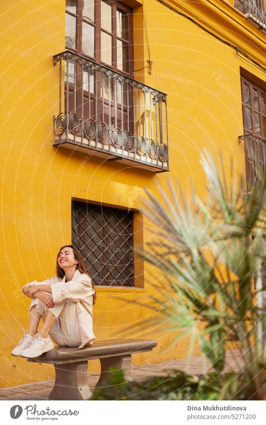 Junge Frau sitzt auf der Bank, lächelnd, in der Nähe von gelben Gebäude mit schönen Balkon. Weiblicher Tourist, der alleine reist. tausendjährig urban Model