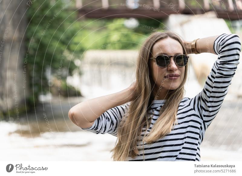 Eine Frau mit Sonnenbrille und gestreiftem Oberteil hält ihr Haar in der Nähe des Flusses mit ihren Händen Top Halt Behaarung Hände. Langarmshirt Trägerkleid