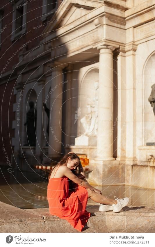 Junge Frau in rotem Kleid sitzt in der Nähe eines Brunnens. Weiblicher Tourist posiert für Fotoshooting. Mode und Stil. rotes Kleid Tourismus reisen im Freien