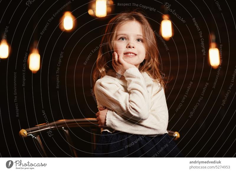Kleine adorable lächelnde Mädchen berührt Gesicht mit ihren Händen und Blick in die Kamera mit einem dunklen Hintergrund. Kind in Spitze blauen Kleid und Strickpullover sitzt auf Metall Stuhl. Glückliche Kindheit