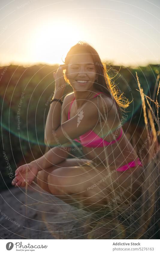 Fröhliche Frau, die am Strand auf den Hüften sitzt Haare berühren Lächeln Sonnenuntergang Sommer Glück Wochenende Urlaub Resort jung sitzen Voute heiter Küste