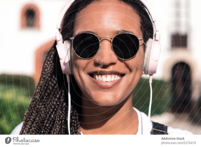 Fröhliche ethnische Frau mit Sonnenbrille und Kopfhörern Lächeln heiter Porträt Musik zuhören Gesang Straße Accessoire Geflecht benutzend Afroamerikaner Stil