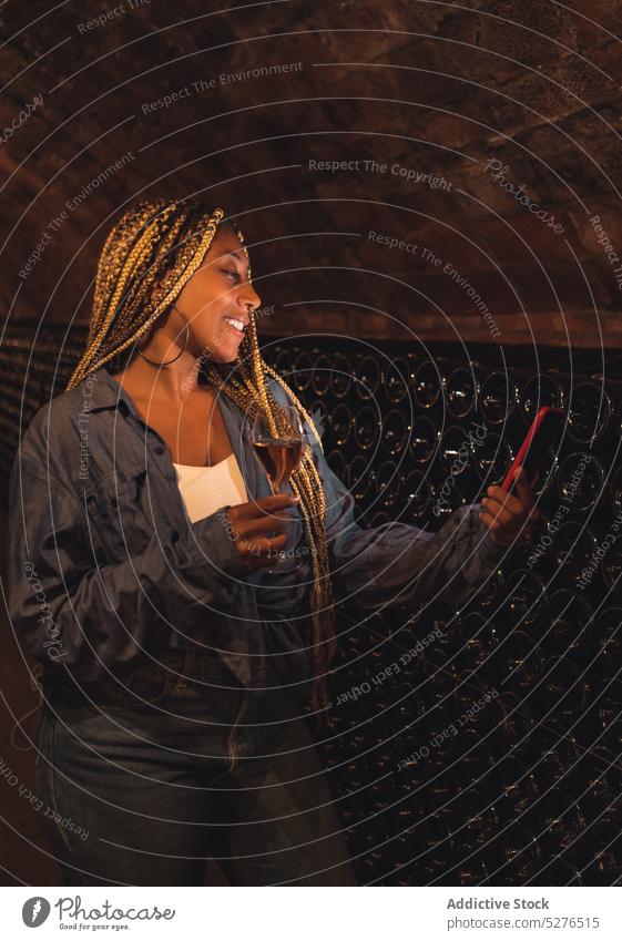 Glückliche junge schwarze Dame mit Weinglas, die ein Videogespräch im Keller führt Frau Smartphone Lächeln degustieren selbstbewusst positiv Alkohol Video-Chat
