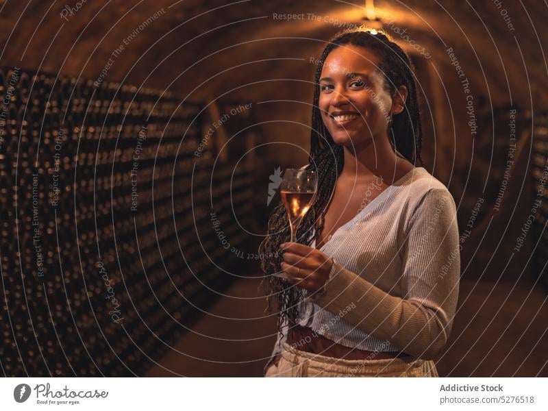 Fröhliche ethnische Dame lächelt beim Weintrinken im Keller Frau Lächeln Alkohol selbstbewusst Weingut genießen Porträt Getränk Flasche jung Afroamerikaner