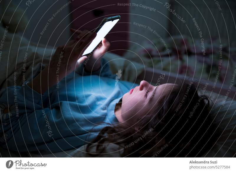 Ein Kind, das spät nachts im Bett liegt und sein Smartphone benutzt, Spiele spielt, Videos online anschaut und auf dem Bildschirm scrollt. Die Bildschirmsucht von Kindern. Das Kinderzimmer bei Nacht.