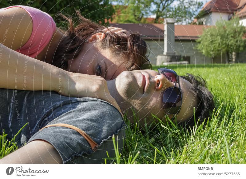 Oberkörper Paar was im Sommer mit T Shirt im  Gras liegt. Sie ist an ihn gekuschelt und blickt in die kamera schaut während er sonnenbrille trägt und mit dem Kopf im Gras liegt.