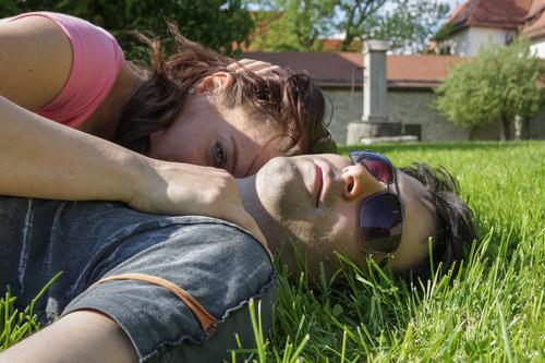 Oberkörper Paar was im Sommer mit T Shirt im  Gras liegt. Sie ist an ihn gekuschelt und blickt in die kamera schaut während er sonnenbrille trägt und mit dem Kopf im Gras liegt.