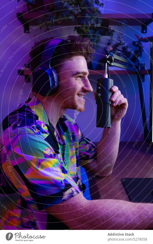 Fröhlicher Mann bei der Aufnahme eines Podcasts im Studio Lächeln heiter Aufzeichnen Mikrofon Kopfhörer neonfarbig unterhalten Atelier Radio jung leuchten Tisch
