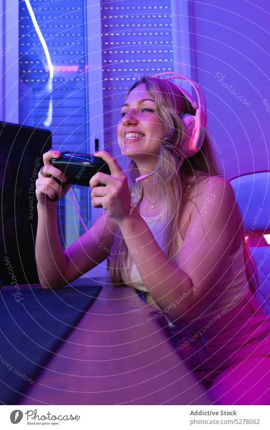 Fröhliche Streamerin mit Kopfhörern, Mikrofon und Gamepad Frau heiter Spiel strömen Konsole spielen Glück Aufzeichnen Blogger neonfarbig Raum Headset reden jung