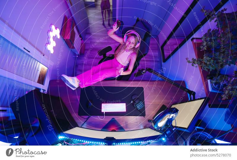 Fröhliche Frau, die in einem dunklen Raum einen Stream aufnimmt heiter Lächeln Blogger Kopfhörer Stuhl Aufzeichnen Atelier strömen Computer neonfarbig modern