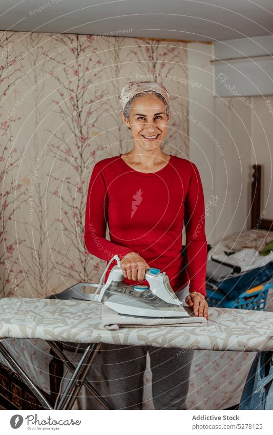 Lächelnde Frau bügelt Kleidung im Zimmer bügeln heimwärts Haushalt Stoff Hausarbeit Routine Arbeit Hausfrau heimisch täglich Appartement Pflicht Kleidungsstück