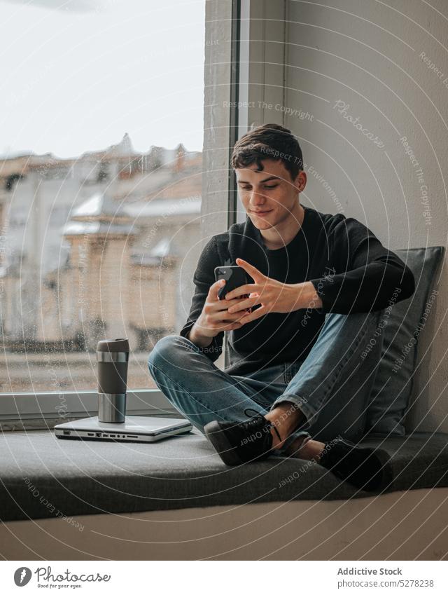 Lächelnder Teenager, der sein Smartphone auf der Fensterbank durchsucht Mann Schüler Pause Laptop heiter Nachricht Fenstersims männlich Beine gekreuzt positiv