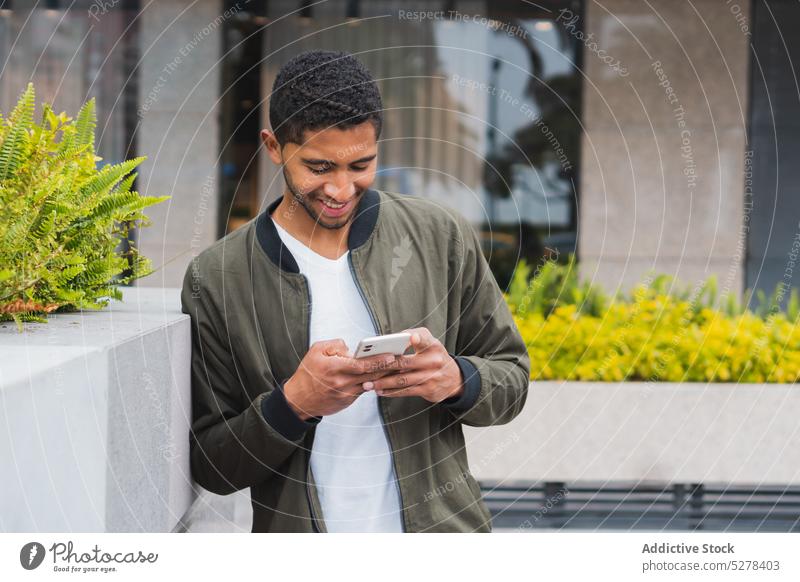 Glücklicher ethnischer Mann, der auf der Straße sein Smartphone benutzt benutzend Lächeln Konzentration Browsen Textnachricht Freude froh Surfen männlich