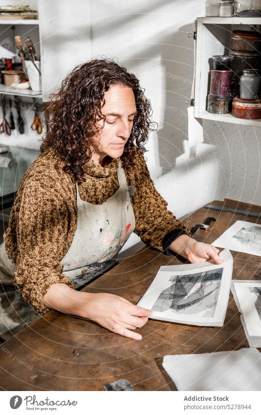 Kunsthandwerkerin bei der Kontrolle gravierter Fotos Handwerkerin untersuchen Fotogravur Papier handgefertigt Tisch Atelier Frau Lebensmitte reif Werkstatt